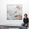 Künstlerin Katja Gramann kniet lächelnd vor ihrem quadratischen Gemälde „Neubeginn Nr. 2" im Atelier