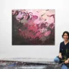 Künstlerin Katja Gramann sitzt lächelnd vor ihrem abstrakten Originalgemälde „Füreinander Nr. 3" in ihrem Atelier in Gräfelfing bei München