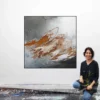 Künstlerin Katja Gramann kniet lächelnd vor ihrem quadratischen Gemälde „Neubeginn Nr. 4" im Atelier